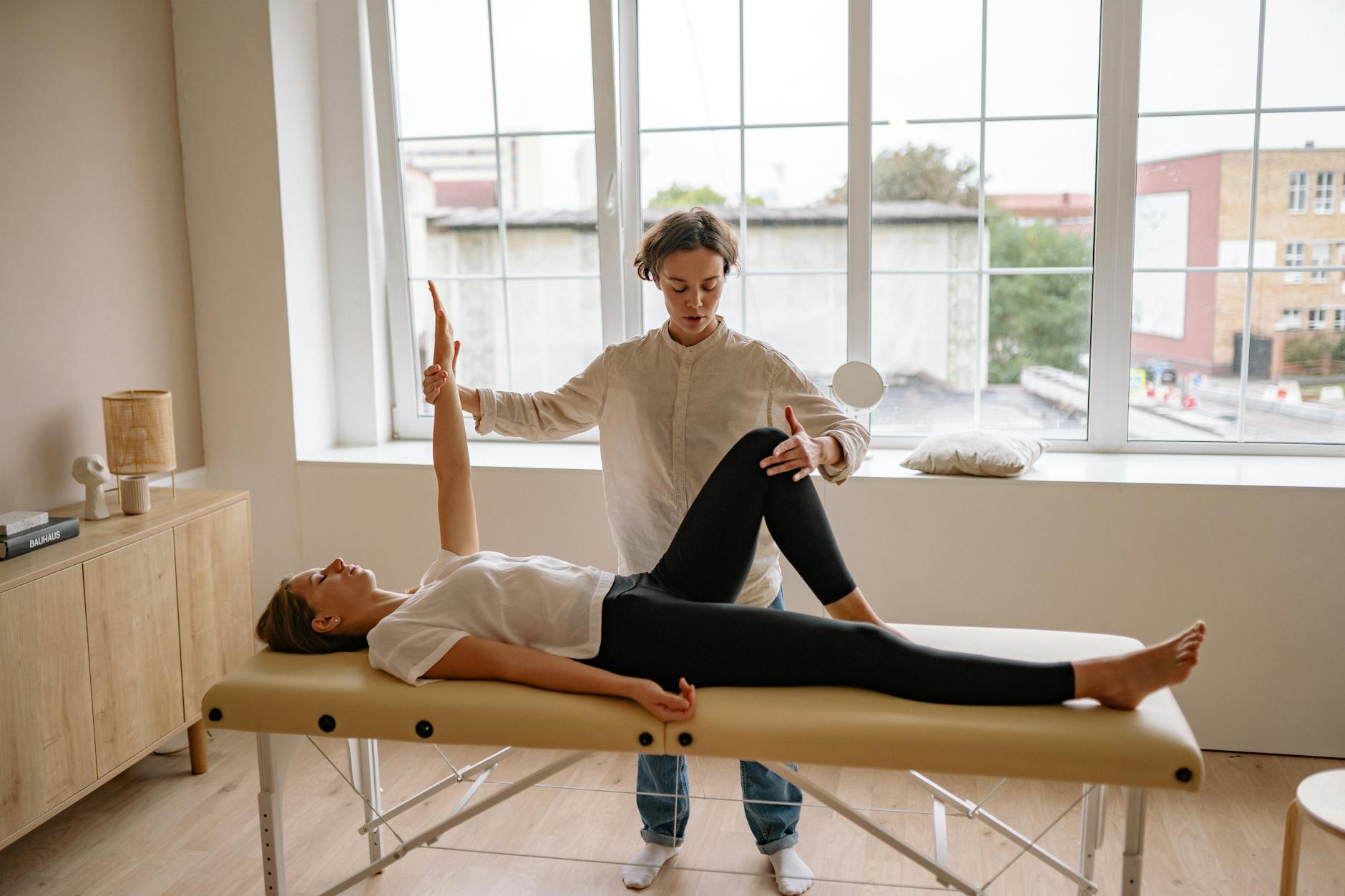 Patient lying on mat while practitioner guides leg stretch in bright room
